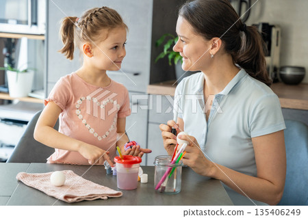 Mother and daughter holding colored eggs while smiling at each other. Shared creativity and bonding during Easter celebration. Mother and daughter holding colored eggs while smiling at each other. Shared creativity and bonding during Easter celebration. 135406249