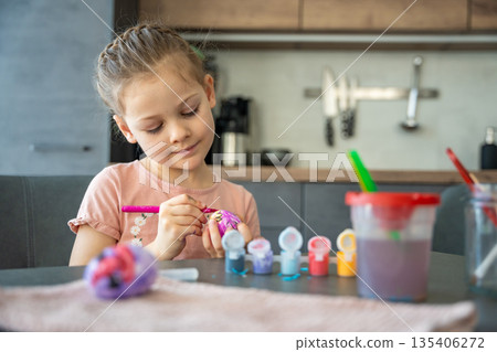 Little girl decorating Easter egg using brush and vivid colors. A moment of childhood joy and focus on holiday traditions. Little girl decorating Easter egg using brush and vivid colors. A moment of childhood joy and focus on holiday traditions. 135406272