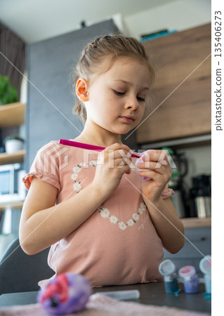 Little girl carefully rotating egg to paint even surface. Home-based creativity and immersive holiday engagement. Little girl carefully rotating egg to paint even surface. Home-based creativity and immersive holiday engagement. 135406273