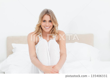 Mid-twenties woman sitting on bed hugging knees in white bedroom wearing pajamas with pillows Mid-twenties woman sitting on bed hugging knees in white bedroom wearing pajamas with pillows 135406331