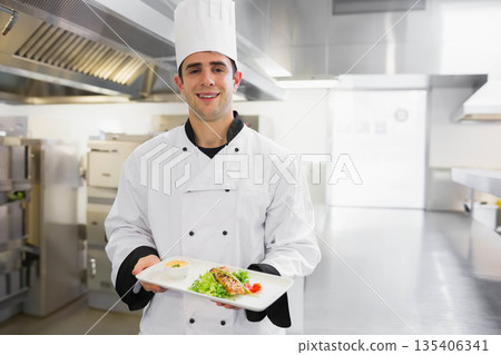 Male chef in chef coat holding plate topped with fish fillet, greens, ramekin in restaurant kitchen Male chef in chef coat holding plate topped with fish fillet, greens, ramekin in restaurant kitchen 135406341