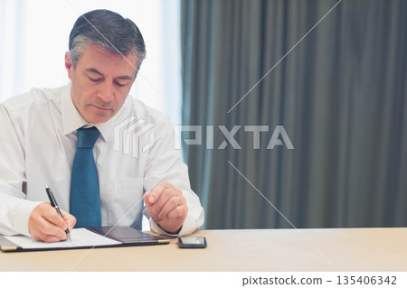 Male wearing white shirt and blue tie signing papers at office table with smartphone, copy space 135406342