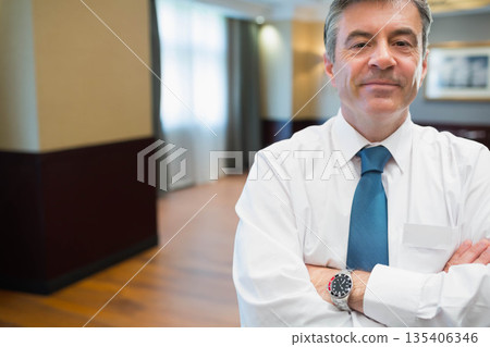 Middle-aged man in shirt and tie crossing arms in hotel corridor featuring wristwatch, copy space 135406346