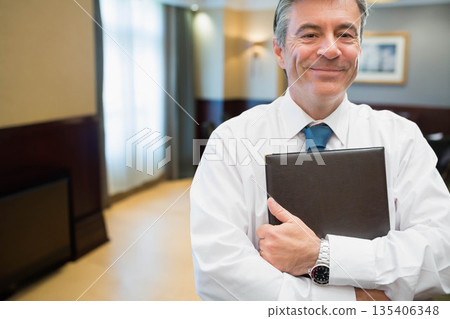 Senior businessman standing in corporate hallway holding binder showing wristwatch, copy space 135406348