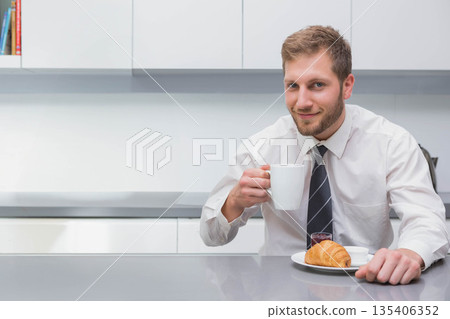 Businessman sitting at countertop in kitchen holding coffee mug and croissant with jam, copy space 135406352