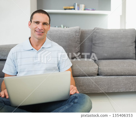Man sitting on floor holding laptop in living room near grey couch with shelf, copy space 135406353