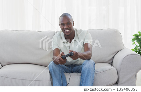 African American man gripping game controller, leaning on couch at home with curtains and plant African American man gripping game controller, leaning on couch at home with curtains and plant 135406355