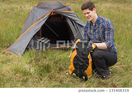 Male camper kneeling at meadow campsite packing hiking backpack by orange dome tent, copy space Male camper kneeling at meadow campsite packing hiking backpack by orange dome tent, copy space 135406359