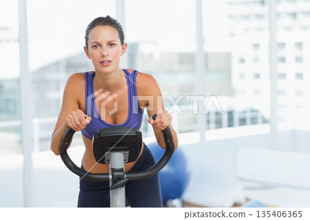 Woman in purple sports top and leggings leaning and gripping bike handlebars at fitness studio 135406365