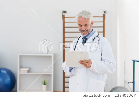 Senior male doctor standing and reading clipboard notes in rehab room with stethoscope, copy space 135406367