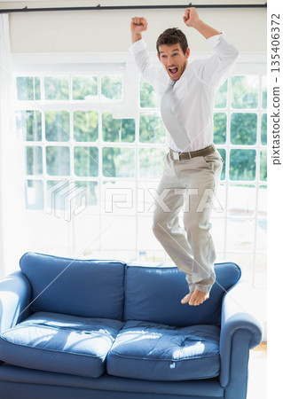 Man in his thirties jumping on blue couch at home under grid windows with curtains 135406372