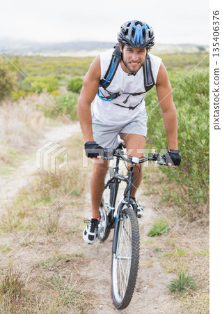 Male cyclist wearing helmet hydration pack gloves riding mountain bike on dirt trail through shrubs 135406376