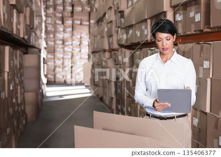 Woman standing in storage aisle holding clipboard and pen inspecting open cardboard box, copy space 135406377