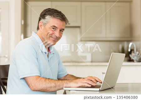 Senior man typing on silver laptop in modern kitchen at table with marble countertop Senior man typing on silver laptop in modern kitchen at table with marble countertop 135406380