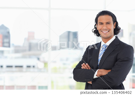 Businessman standing with arms crossed by office lobby windows revealing city skyline, copy space Businessman standing with arms crossed by office lobby windows revealing city skyline, copy space 135406402