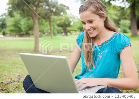 Woman working on silver laptop while sitting cross-legged in city park wearing turquoise blouse Woman working on silver laptop while sitting cross-legged in city park wearing turquoise blouse 135406403