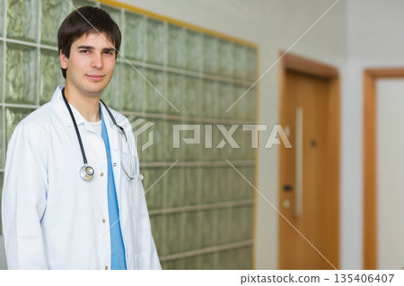 Male doctor standing in hospital corridor wearing white lab coat blue scrubs and stethoscope Male doctor standing in hospital corridor wearing white lab coat blue scrubs and stethoscope 135406407
