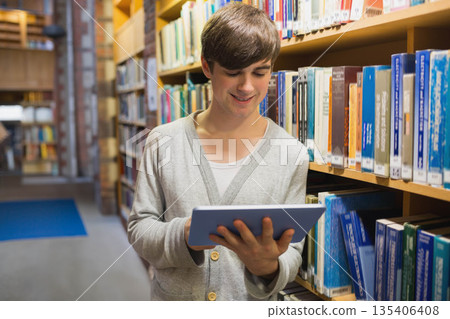 Male student holding tablet and reading info while standing in library aisle with bookshelves Male student holding tablet and reading info while standing in library aisle with bookshelves 135406408