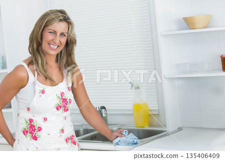 Woman wearing apron cleaning sink at kitchen countertop with cloth and spray bottle, copy space 135406409