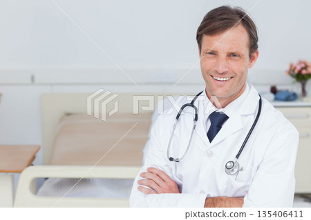 Smiling male doctor wearing lab coat and stethoscope standing in hospital room near bed, copy space 135406411