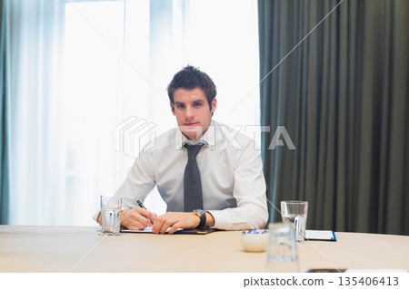 Businessman writing notes on notepad at meeting table under sheer curtains with water glasses Businessman writing notes on notepad at meeting table under sheer curtains with water glasses 135406413