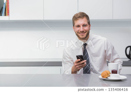 Male in suit holding smartphone and smiling at kitchen counter with mug, croissant, copy space Male in suit holding smartphone and smiling at kitchen counter with mug, croissant, copy space 135406416