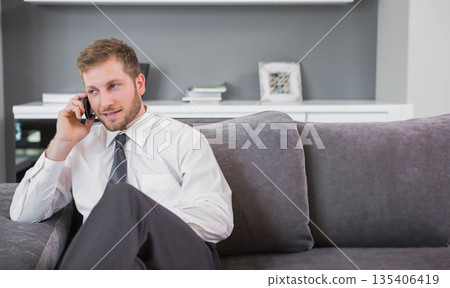 Man in business attire calling on smartphone, sitting on grey sofa near white console, copy space 135406419