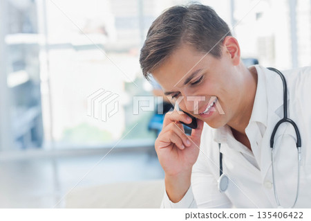 Male doctor wearing lab coat smiling while talking on smartphone by window in clinic, stethoscope Male doctor wearing lab coat smiling while talking on smartphone by window in clinic, stethoscope 135406422