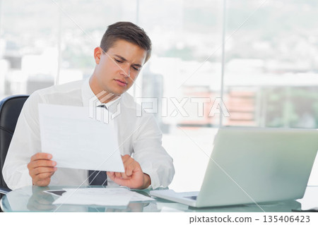 Male office worker in suit holding document, glancing at wristwatch at desk with laptop in office 135406423