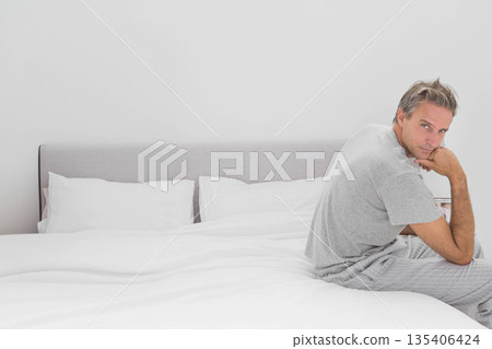 Middle-aged man sitting on bed in bedroom wearing grey T-shirt and striped pajama pants, copy space Middle-aged man sitting on bed in bedroom wearing grey T-shirt and striped pajama pants, copy space 135406424