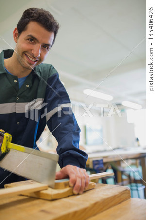 Man wearing reflective coverall sawing wooden plank with yellow-handled saw at workshop, copy space 135406426