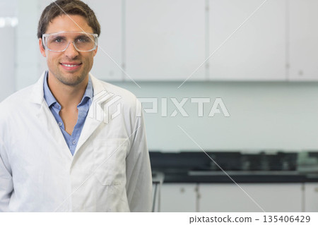 Man wearing white lab coat, goggles standing in lab by countertop with cabinet drawers, copy space Man wearing white lab coat, goggles standing in lab by countertop with cabinet drawers, copy space 135406429