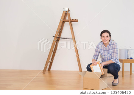 Woman kneeling in room unpacking frame from cardboard box beside ladder and paint cans, copy space 135406430