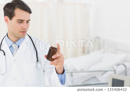Male doctor holding amber bottle, examining label with stethoscope beside bed in clinic, copy space Male doctor holding amber bottle, examining label with stethoscope beside bed in clinic, copy space 135406432
