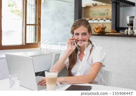 Smiling woman holding smartphone to ear while typing on laptop at cafe table with latte 135406447