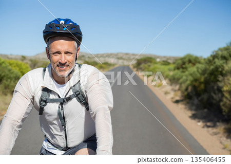 Male cyclist gripping handlebars riding on paved road wearing blue helmet with backpack, copy space Male cyclist gripping handlebars riding on paved road wearing blue helmet with backpack, copy space 135406455