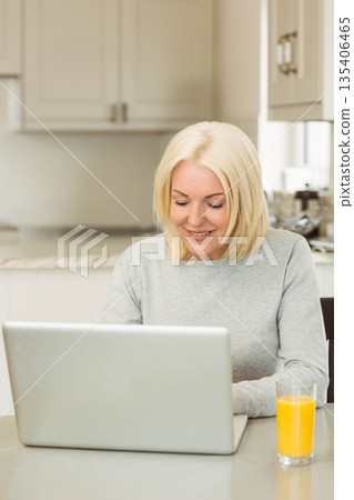 Woman working on laptop at kitchen table with glass of orange juice by coffee machine Woman working on laptop at kitchen table with glass of orange juice by coffee machine 135406465
