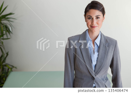 Businesswoman standing in office corner wearing suit beside mint-green desk and plant, copy space 135406474