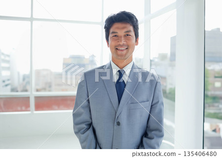 Japanese man wearing grey suit and blue tie standing in bright office with floor-to-ceiling windows Japanese man wearing grey suit and blue tie standing in bright office with floor-to-ceiling windows 135406480