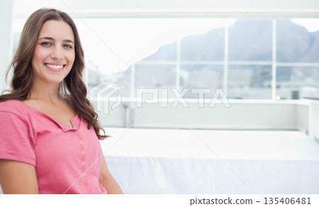 Woman sitting on white bed edge in sunlit glass room overlooking mountain terrace, copy space 135406481