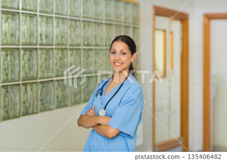 Female nurse standing in hospital corridor wearing blue scrubs, stethoscope, crossing arms, smiling 135406482