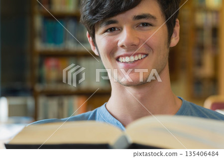 Male student sitting at study table holding large open book in library with bookshelves, copy space Male student sitting at study table holding large open book in library with bookshelves, copy space 135406484