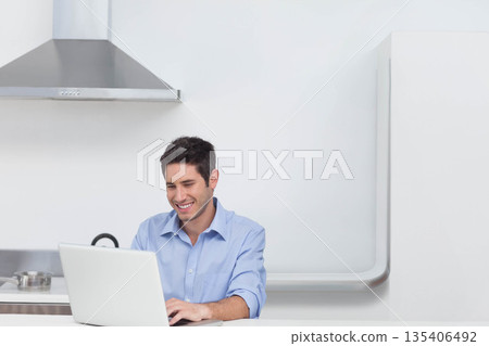 Man typing on silver laptop at white countertop in minimalist kitchen with pot simmering 135406492