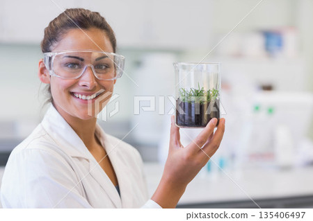 Female scientist wearing lab coat holding glass beaker with soil and seedlings at lab bench Female scientist wearing lab coat holding glass beaker with soil and seedlings at lab bench 135406497