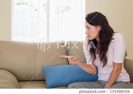 Woman pointing left sitting on sofa in living room with blue cushion near window, copy space 135406506