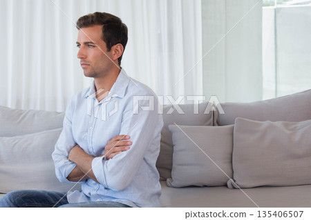 Man in his thirties sitting on light-gray sofa under white sheer curtain in bright living room Man in his thirties sitting on light-gray sofa under white sheer curtain in bright living room 135406507