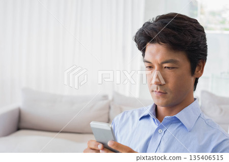 Mid adult Asian man sitting on sofa at home holding smartphone wearing blue shirt, copy space 135406515