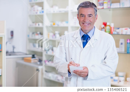 Middle aged male pharmacist writing on notepad with pen at pharmacy desk with shelves, copy space Middle aged male pharmacist writing on notepad with pen at pharmacy desk with shelves, copy space 135406521