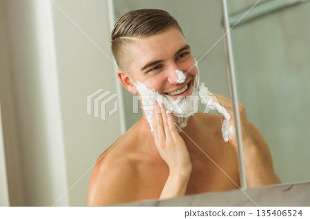 Shirtless man spreading thick shaving foam across face and cheeks in modern bathroom mirror Shirtless man spreading thick shaving foam across face and cheeks in modern bathroom mirror 135406524