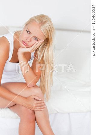 Woman in white top, shorts sitting on edge of bed in bedroom, resting chin on hand Woman in white top, shorts sitting on edge of bed in bedroom, resting chin on hand 135406534